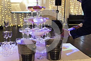 Waiter pours the champagne into the glasses tower at the illuminated banquet hall background. The glasses pyramid with sparkling