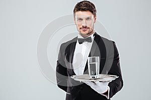 Waiter in tuxedo holding glass of water on metal tray