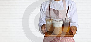 Waiter wearing pink apron carries 3 take away cups of ice coffee for serving on white brick background with copy space