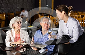 Waitress bringing wine to senior couple choosing dishes from menu