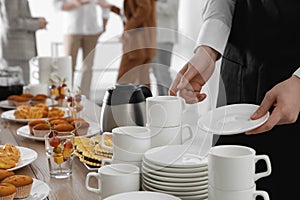 Waitress near table with dishware and different delicious snacks during coffee break, closeup