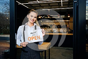 Waitress wearing apron standing at restaurant entrance. Woman attend new customers in her coffee shop. Happy woman showing open photo