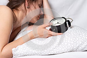 Wake up call. Young beautiful woman lying in bed and touching alarm clock. Girl doesn`t want to wake up