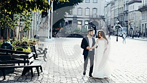 Walk of the happy smiling newlywed couple actively talking and holding hands. Sunny street town location.