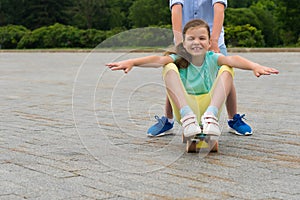 On a walk in the park, the boy pushes a girl in front of him, who sits on a skateboard