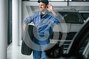 Walking forward and holding the tire. Man in blue uniform is working in the car service