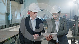 Walking in front of the camera in manufacturing factory two engineers and analysing the plan of production while holding