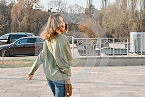 Walking young smiling girl teenager looking in camera through back with brown red hair in green sweater, sunny spring day