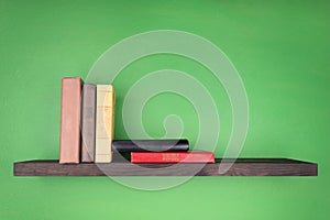 On the wall of green color there is a dark wooden shelf with a texture on which several books stand vertically from the left and
