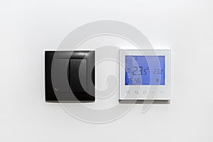 A wall-mounted room thermostat showing a temperature of 23.5 degrees Celsius, next to it a black switch for roller blinds.