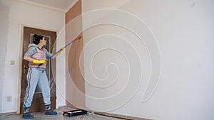 wall painting. a woman, in work clothes, paints a white wall indoors with pink paint using a roller on a long holder.