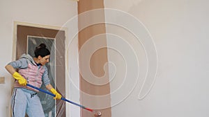 wall painting. a woman, in work clothes, paints a white wall indoors with pink paint using a roller on a long holder.