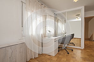 Wall of a room with a white wooden desk with swivel chair next to a built-in cupboard with sliding glass doors with white edges