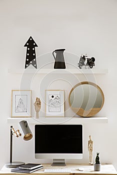 Wall shelves with decor and simple posters above wooden home office desk with metal lamp, mockup monitor and notebooks in white in