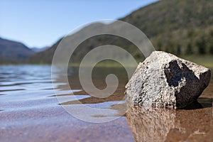 Wallpaper of isolated gray strone in transparent lake water with a huge mountain with forests and a clear sky in Lago Falkner,