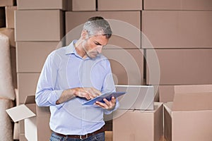 Mid-adult man wearing blue shirt, jeans working on tablet among boxes with laptop in warehouse