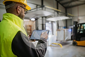 Warehouse receiver standing by delivered cargo, holding tablet, looking at cargo details, checking delivered items