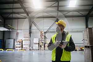 Warehouse receiver standing by delivered cargo, holding tablet, looking at cargo details, checking delivered items