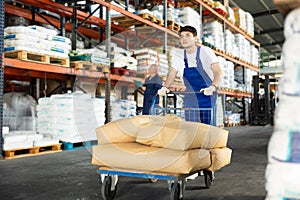 In warehouse of store, stevedore loader young man pushes and carries large cart for bulky cargo