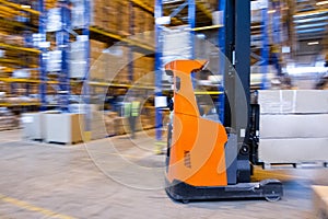 Warehouse worker driving a forklift truck full of goods between high blue yellow steel shelves inside warehouse