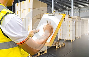 Warehouse worker holding clipboard writing on paper checklist for delivering shipment boxes on pallets.