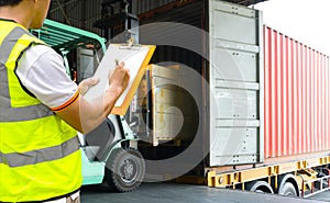 Warehouse worker holding clipboards control forklift loading shipment goods into container truck.