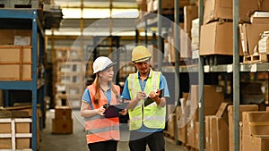 Warehouse workers wearing safety hardhat discussing work while walking in aisle between rows of tall shelves full of