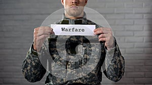 Warfare word written on sign in hands of male soldier, military conflict