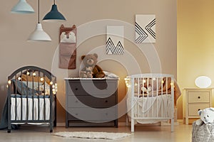 Warm baby bedroom interior with white and grey cribs, commode and small nightstand table with lamp