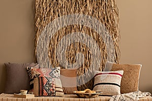 Warm composition of living room interior with dried straw on wall, couch, colorful pillows, carlet plaid, brown wall, book, cup,