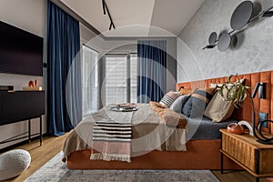 Warm and cozy bedroom interior with big orange bed, beige and grey bedclothes, blue curtain, tv, wooden night stand, black consola