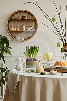 Warm and cozy composition of easter dining room interior with round table, cake, hare sculpture, colorful eggs, shelf on wall,