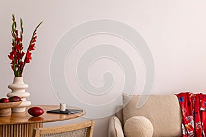 Warm and cozy interior of living room space with round wooden table, beige sofa, red flowers, kimono, rattan chair, decoration.