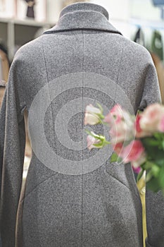 A warm grey coat close up is dressed on a mannequin in clothing store. In the foreground is bouquet of flowers