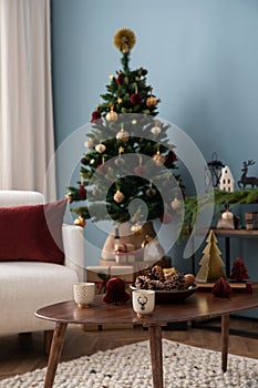 Warm and stylish christmas living room interior with design armchair, wooden coffee table, christmas tree, balls, gifts, red