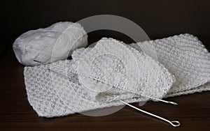 Warm white knitted scarf, knitting needles and a skein of wool yarn on a dark background