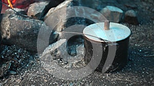 Warming up water in a metal saucepan next to a bonfire outdoors during a hike with burning wooden logs and smoke