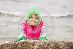 Warmly dressed girl looks out from behind a rock against the backdrop of the sea on cold cloudy day