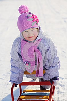 Warmly dressed smiling girl in pink scarf and hat relies on sled