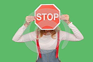 Warning of ban, no access. Portrait of anonymous woman in stylish overalls covering face with octagonal red Stop traffic sign