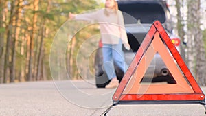 Warning red triangle. Sad young woman sits in trunk of gray car with flashing emergency light along road waiting for