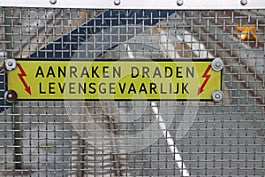 Warning sign with dutch text `aanrakeen draden levensgevaarlijk; which means touching wires is deadly, above electricity wires of