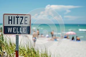 Warning sign with German text \'Hitzewelle\' (Heat wave) in front of blurry sunny beach with people