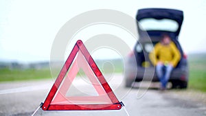 Warning triangle sign on road with man in blur background sitting in trunk of broken car