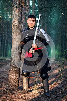 Warrior in black armor with a sword in his hands is leaning against a tree in a gloomy enchanted, damned forest