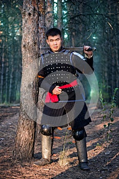 Warrior in black armor with a sword in his hands is leaning against a tree in a gloomy enchanted, damned forest photo