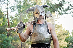warrior in a horned helmet, a steel breastplate with a two-handed ax in his hands, posing against the backdrop of the forest