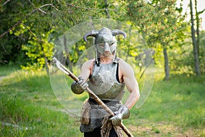 warrior in a horned helmet, a steel breastplate with a two-handed ax in his hands, posing against the backdrop of the forest