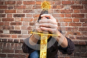 Warrior kneels in a hooded cloak and holds a shining golden sword in front of him with both hands. Knight swears allegiance.