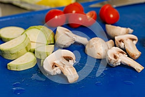 Washed and cut raw mushrooms, zucchini wand cherry tomatoes on blue plastic cutting board - Raw food in a real kitchen concept.